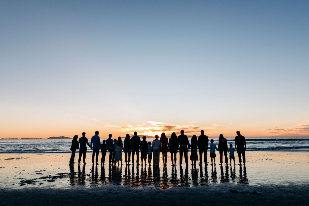 Silhouettes of a large group of people standing together against a dramatic sunset sky at the beach.