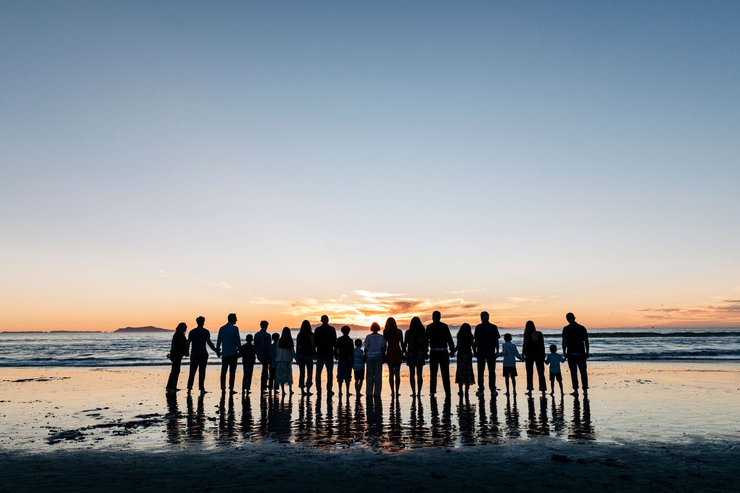 Silhouettes of a large group of people standing together against a dramatic sunset sky at the beach.