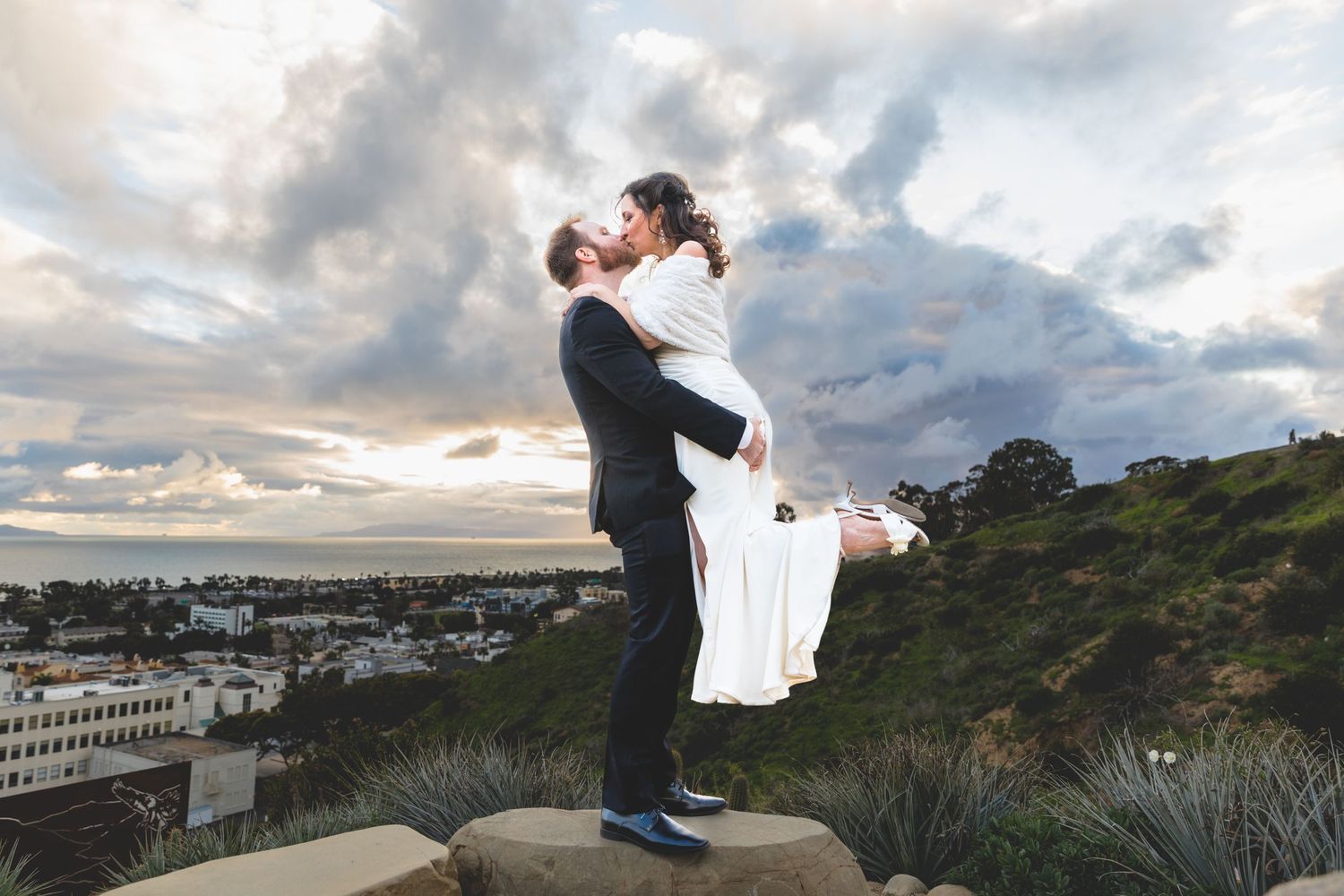 Romantic sunset portrait of couple embracing on clifftop overlooking city.