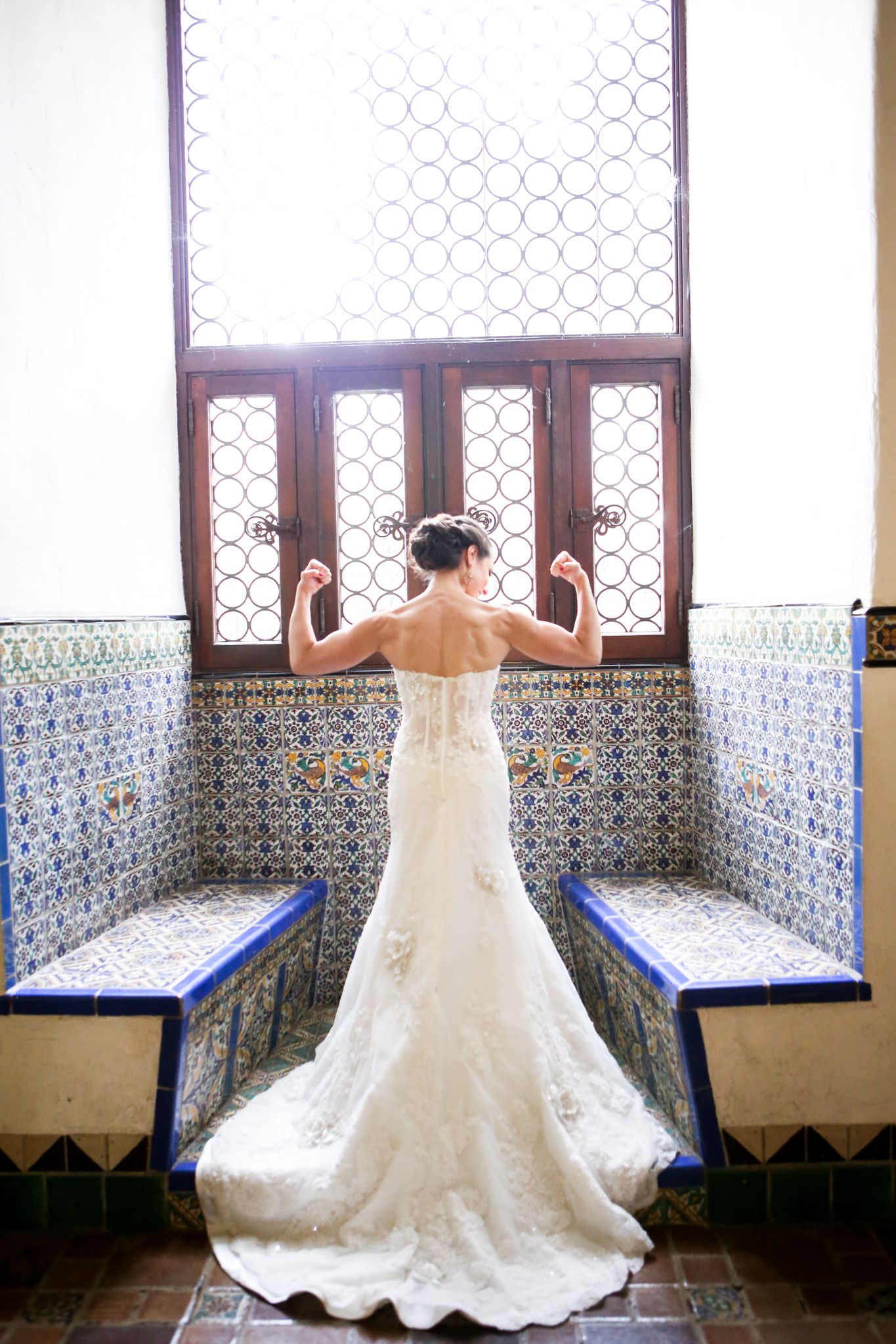 A bride in a flowing white lace gown poses gracefully in a Spanish-style hallway with ornate tile work.