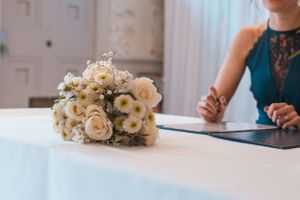 A delicate white and blush rose wedding bouquet rests on a white tablecloth next to someone in a teal dress.
