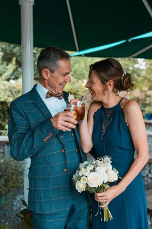 A couple shares a joyful moment under a tent while holding drinks and a white bouquet.
