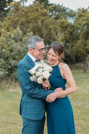 A couple embraces outdoors while holding a white floral bouquet against a natural backdrop.