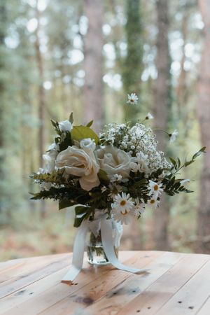 White roses and daisies arranged in a glass vase on a rustic wooden table with forest backdrop.