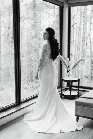 Elegant white wedding dress flowing against large windows with forest view.