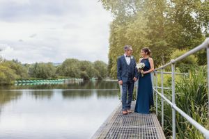 A couple stands on a lakeside dock surrounded by trees while holding a wedding bouquet.