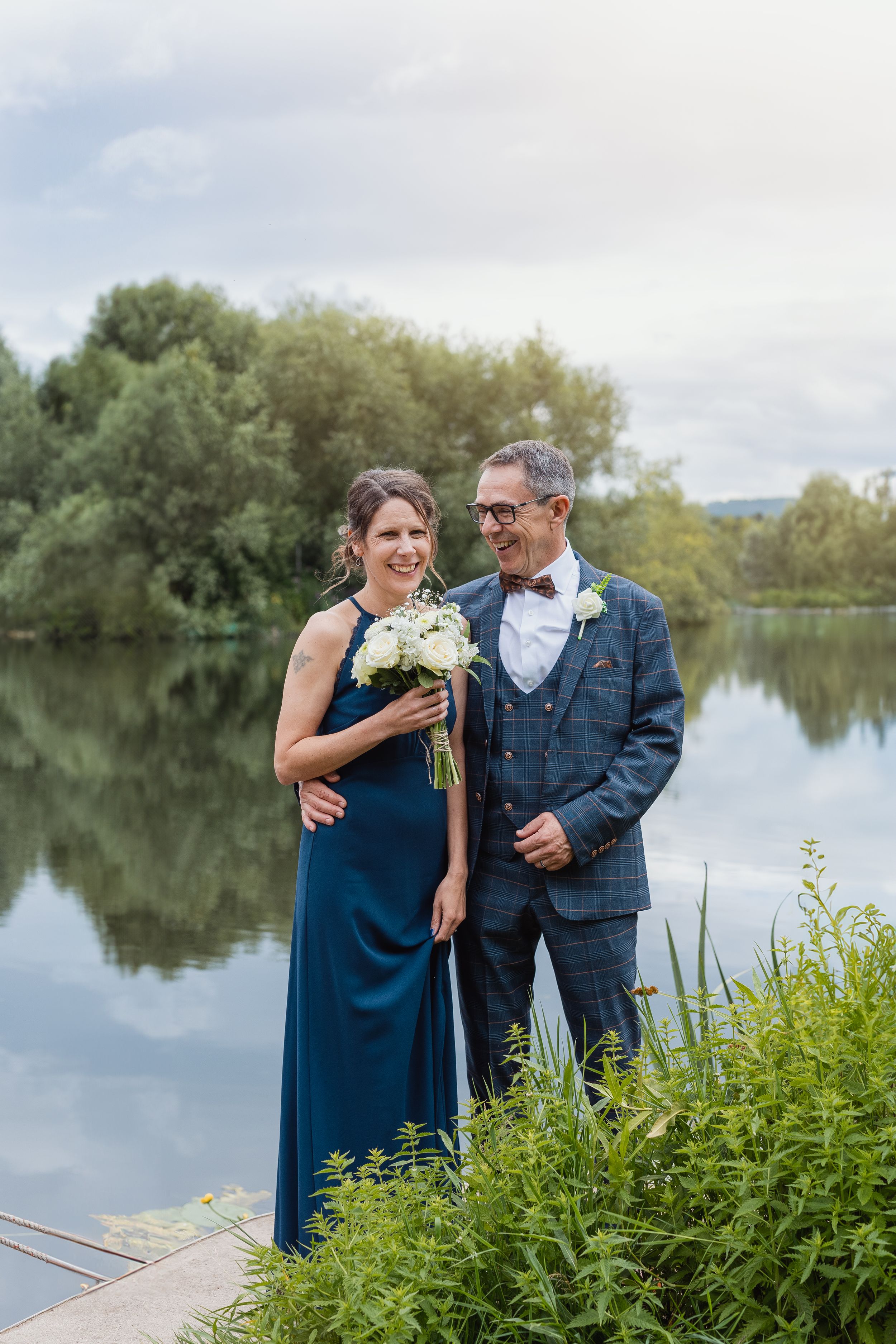 A couple poses by a serene lake with lush greenery, holding a white floral bouquet.