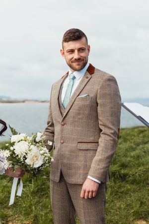 A groom in a brown tweed suit smiles while standing on green coastal cliffs.