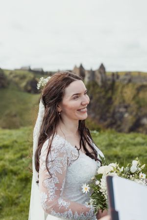 A bride in a lace white dress holds a bouquet against a scenic coastal backdrop.