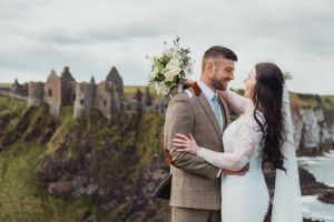 Newlyweds embrace on a cliff overlooking historic castle ruins and ocean views.