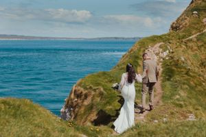 A wedding couple walks along a dramatic coastal path with turquoise ocean views.