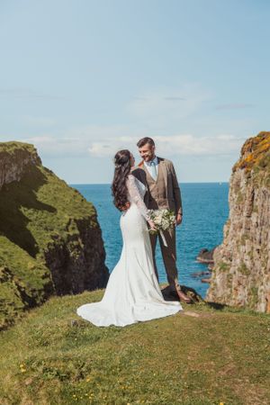 Wedding couple sits on grassy cliff overlooking dramatic ocean coastline on sunny day.