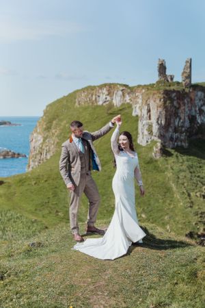 Newlyweds dancing and twirling on a grassy clifftop overlooking the ocean.