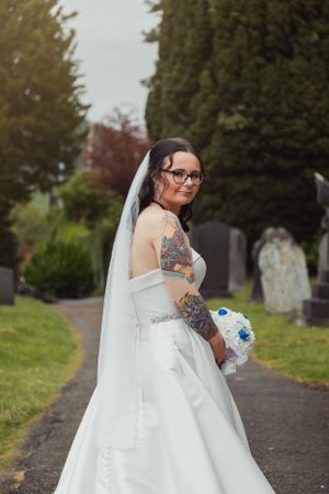 Bride in white dress and veil poses outdoors in a cemetery path surrounded by autumn trees.