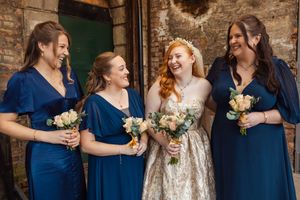 Bridesmaids in navy blue dresses hold white bouquets while laughing and smiling at a rustic brick venue.