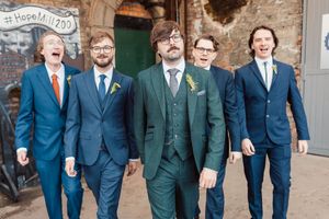 Groomsmen in matching navy blue suits laugh together while standing outside a rustic stone building.