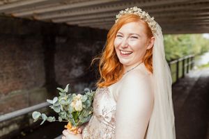 A bride in a lace wedding dress with long red hair holds a eucalyptus bouquet while smiling under a bridge.
