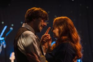 Two performers share an intimate moment on stage with dramatic lighting in the background.