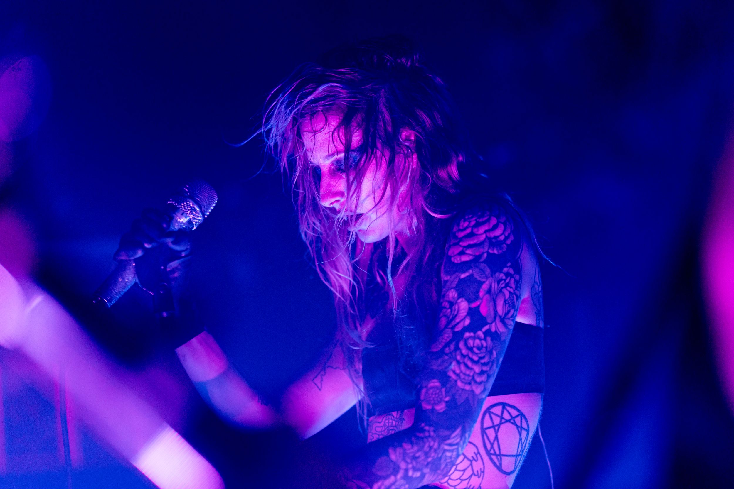 A performer with tattoos sings into a microphone against dramatic blue stage lighting during a concert.