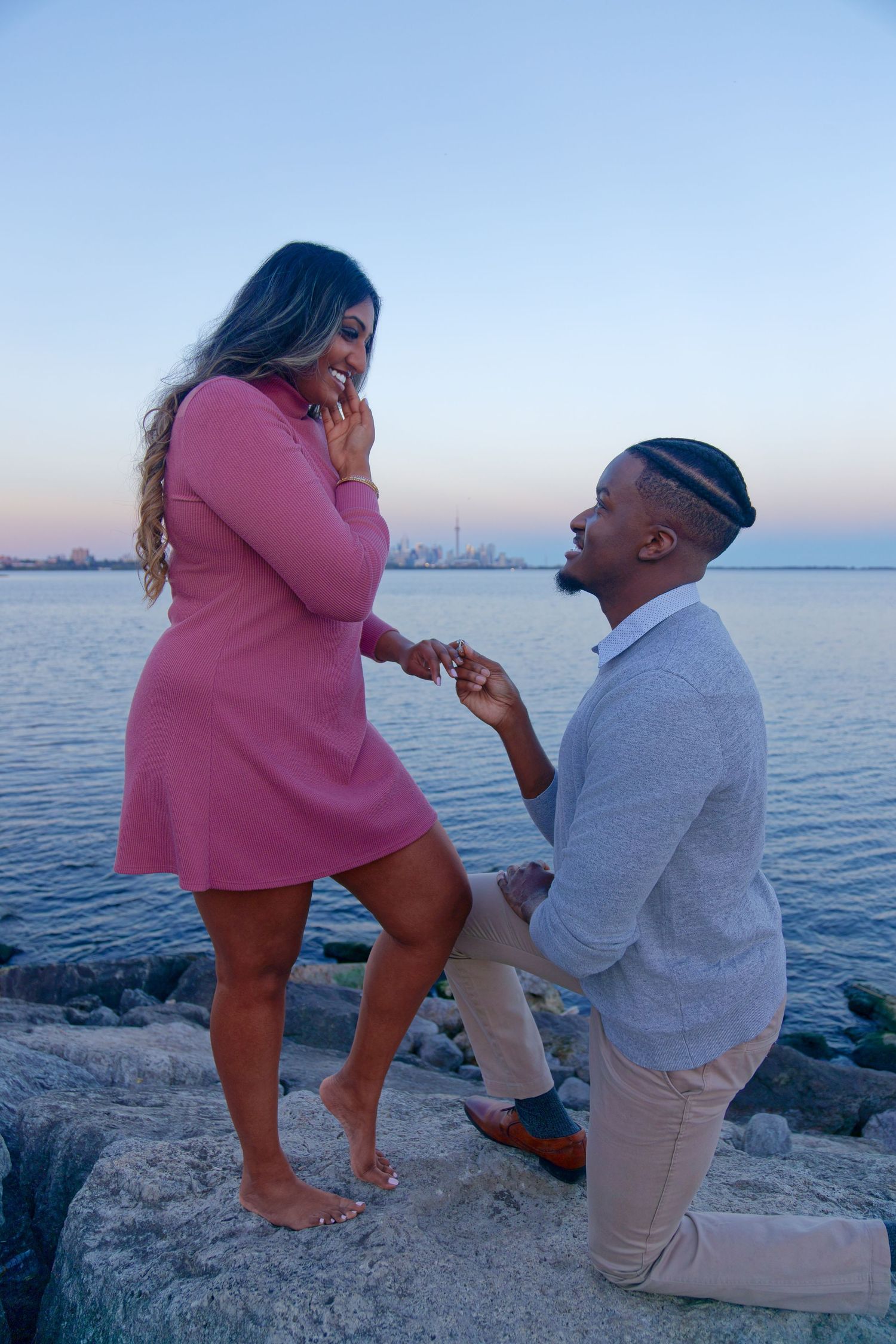 Man proposes to woman in pink dress on rocky waterfront at dusk, city skyline in background.