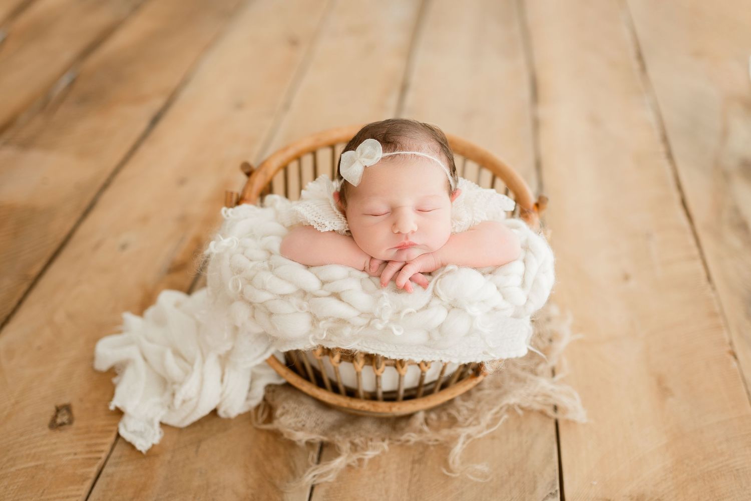 Sleeping newborn wrapped in soft white fabric resting in wooden bowl on hardwood floor.