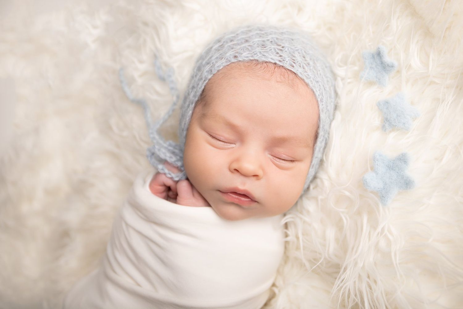 Sleeping newborn wearing blue knit hat rests on cream colored fuzzy blanket.