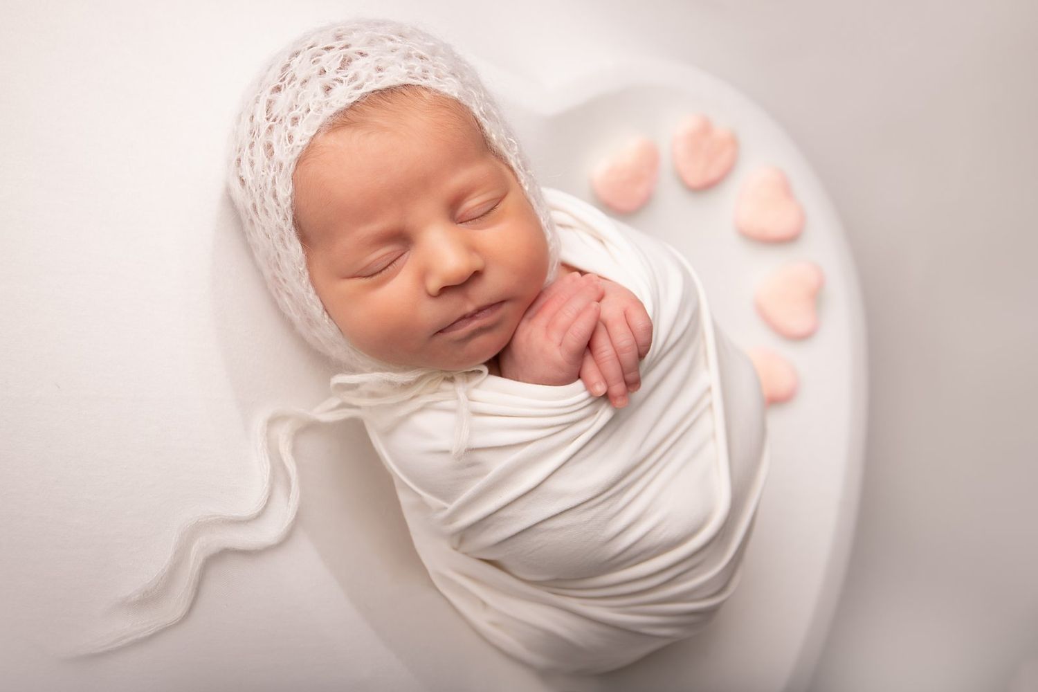 Peaceful sleeping newborn baby wrapped in white swaddle with lace bonnet on white background with pink heart decorations.