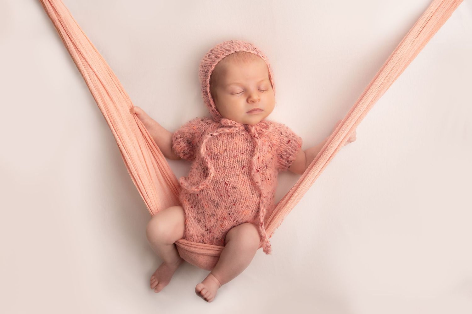 Sleeping newborn in pink knit outfit rests in peach-colored fabric hammock.