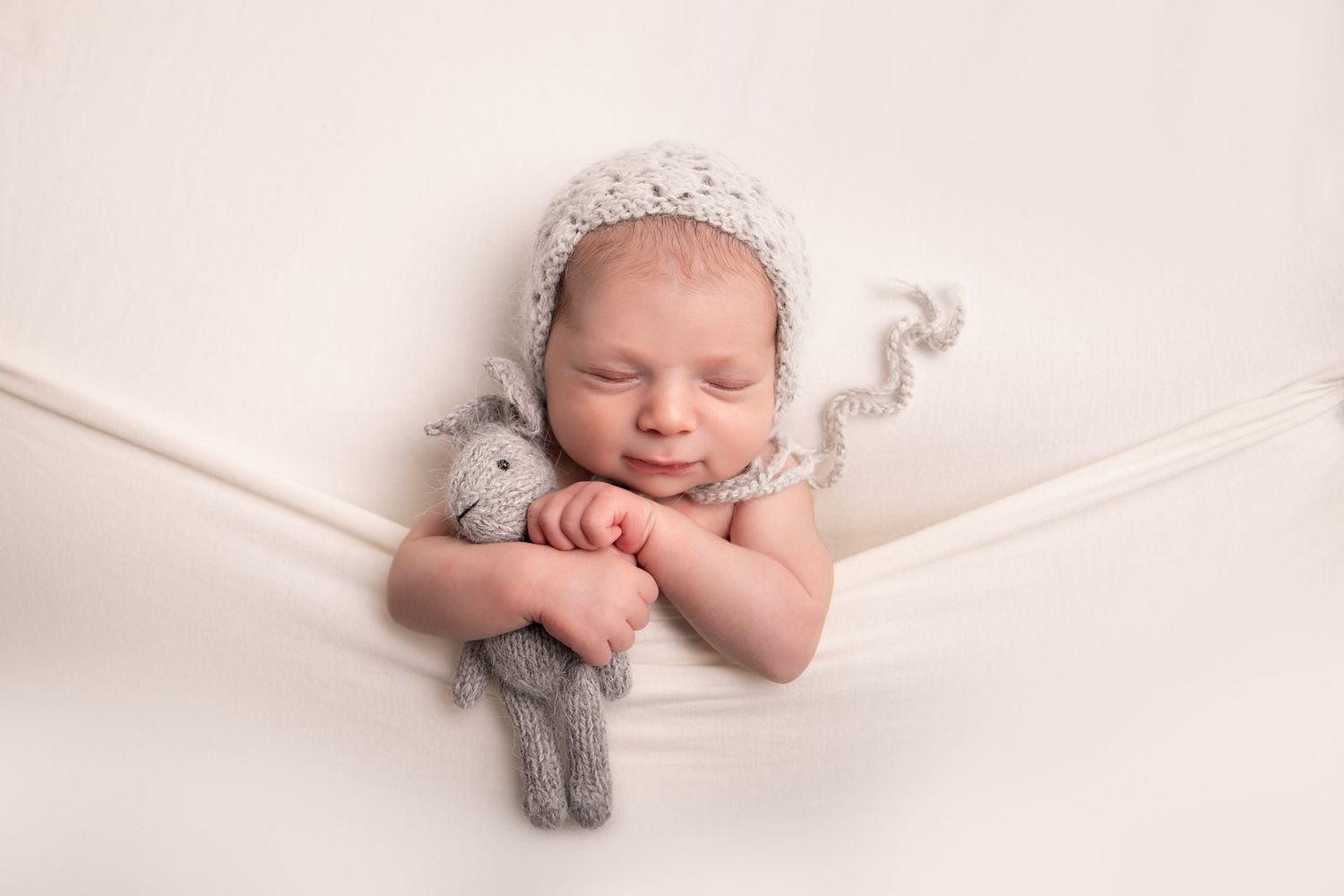 A sleeping newborn baby wearing a knit bonnet lies on a white hammock while holding a small stuffed toy.