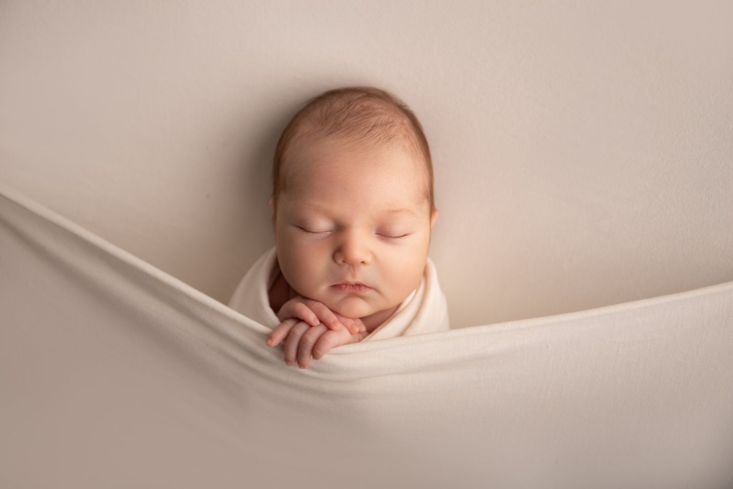 A peaceful sleeping newborn baby nestled in soft beige fabric with their tiny hand resting near their face.