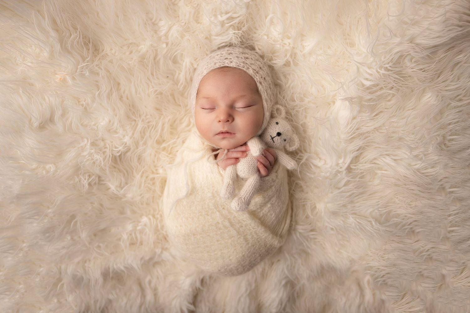 A sleeping newborn wrapped in a cream colored swaddle rests peacefully on a soft fluffy white fur blanket.