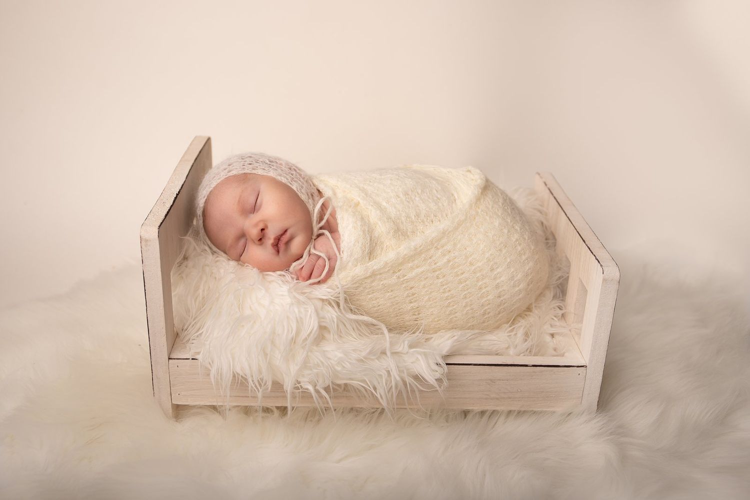 A sleeping newborn wrapped in cream fabric lies in a small wooden bed lined with white fur.