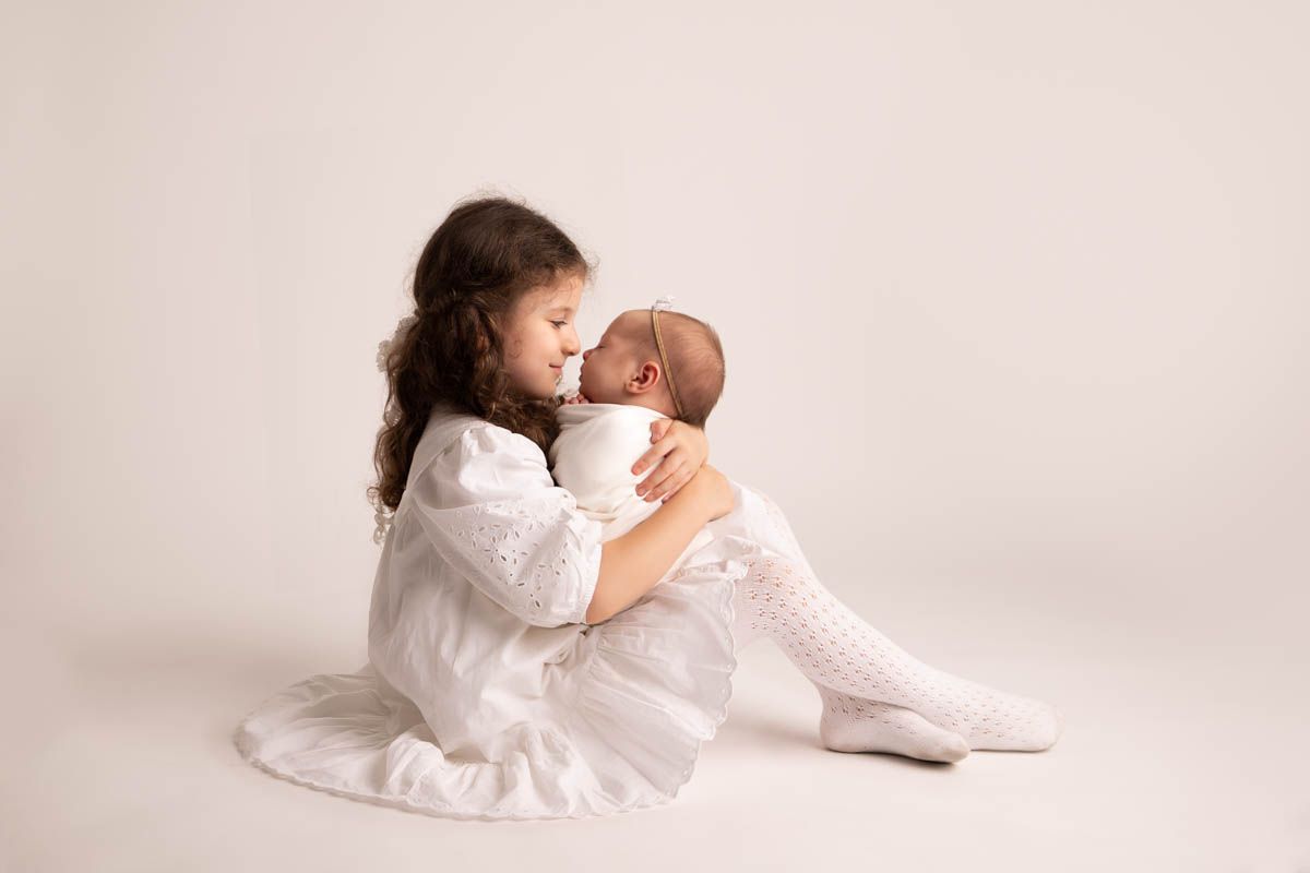 Child in white dress holds newborn while sitting against white studio background.