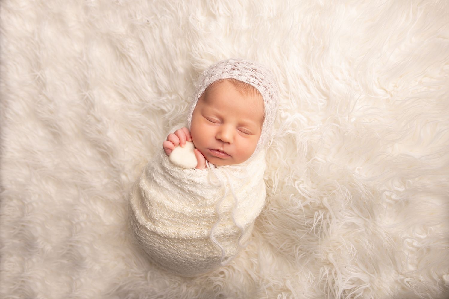 Sleeping newborn wrapped in white knit fabric on fluffy cream background.