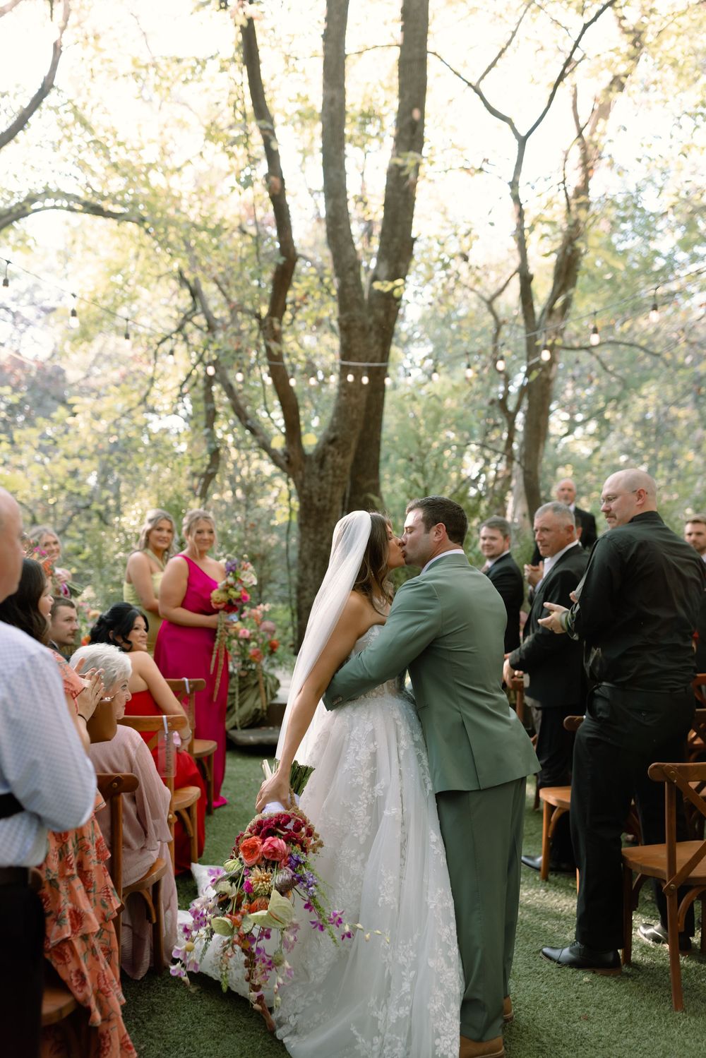 A romantic outdoor wedding ceremony in a wooded setting with guests watching as the couple shares their first kiss.