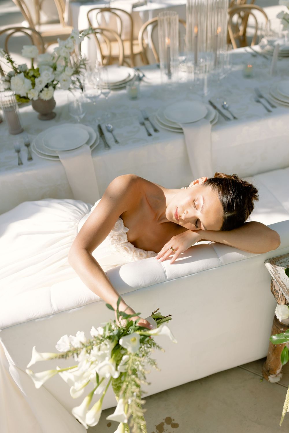 A white chaise lounge with floral arrangements in a bright sunlit room with elegant table settings in the background.