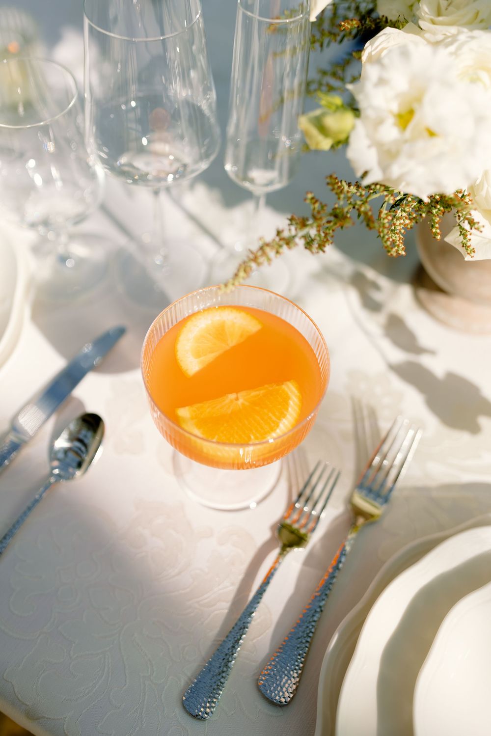 An orange cocktail with citrus garnish sits on a white tablecloth next to elegant place settings and wine glasses.