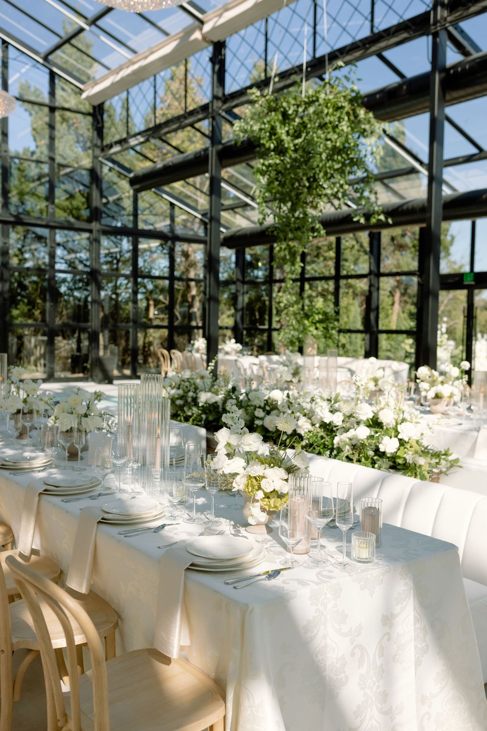 Elegant greenhouse wedding reception setup with white tablecloths, floral centerpieces and natural light streaming through glass ceiling.