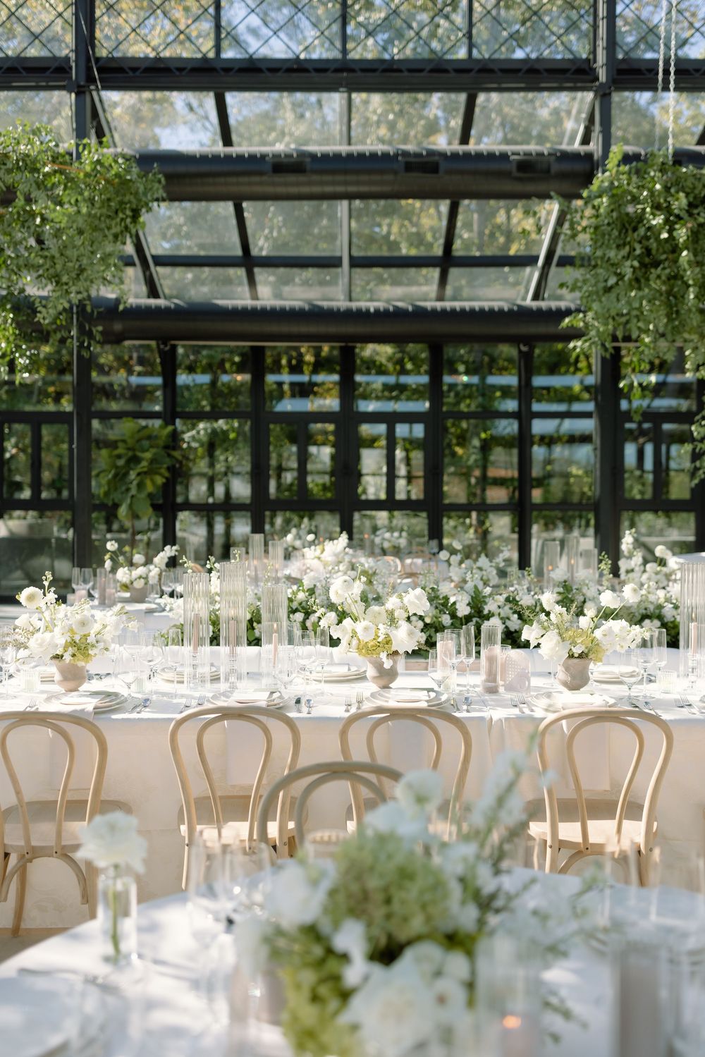 Elegant wedding reception setup with white tables, chairs and floral centerpieces in a bright greenhouse-style venue.