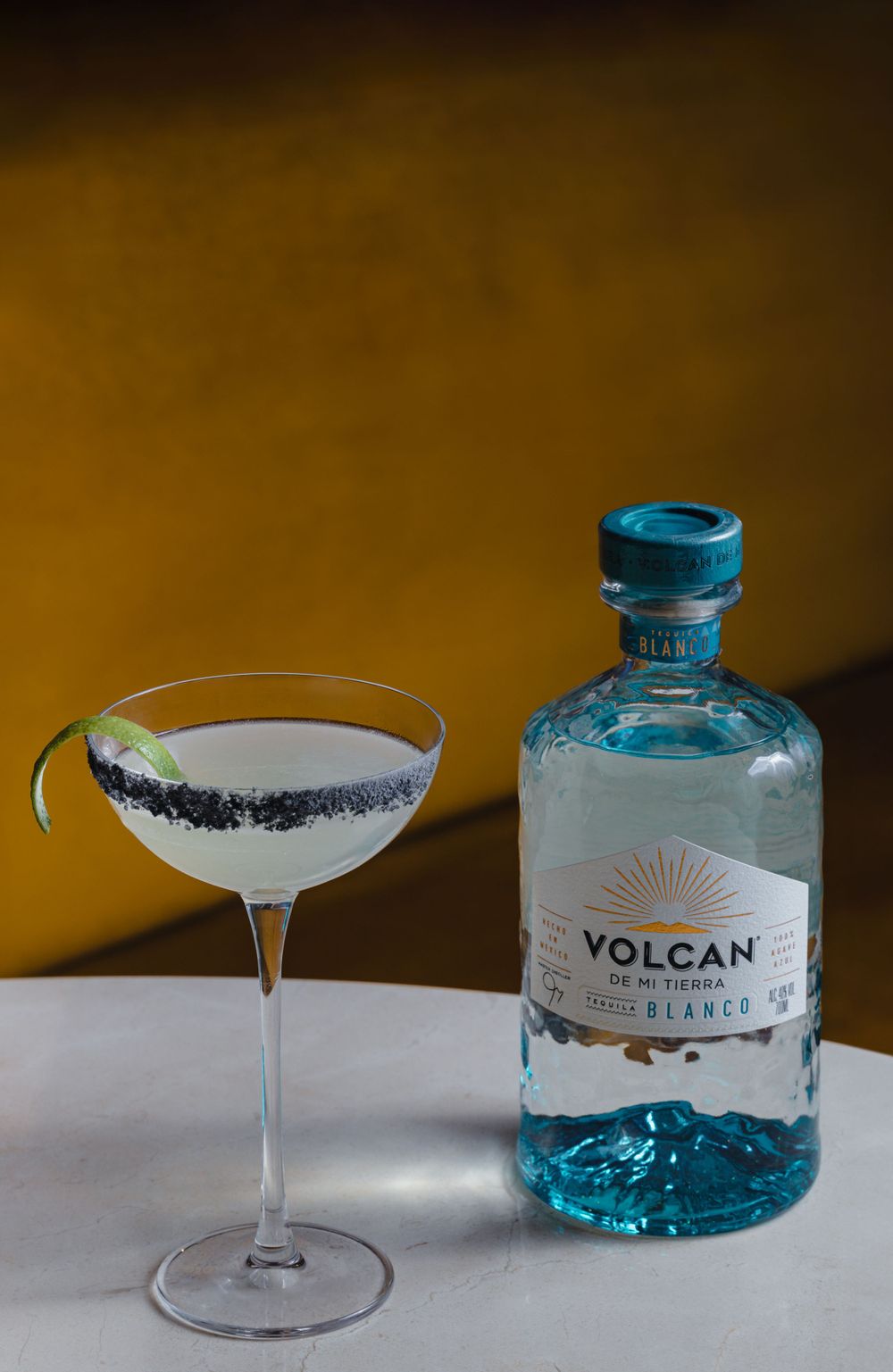 Volcan tequila bottle beside coupe glass with black-rimmed cocktail against golden background.
