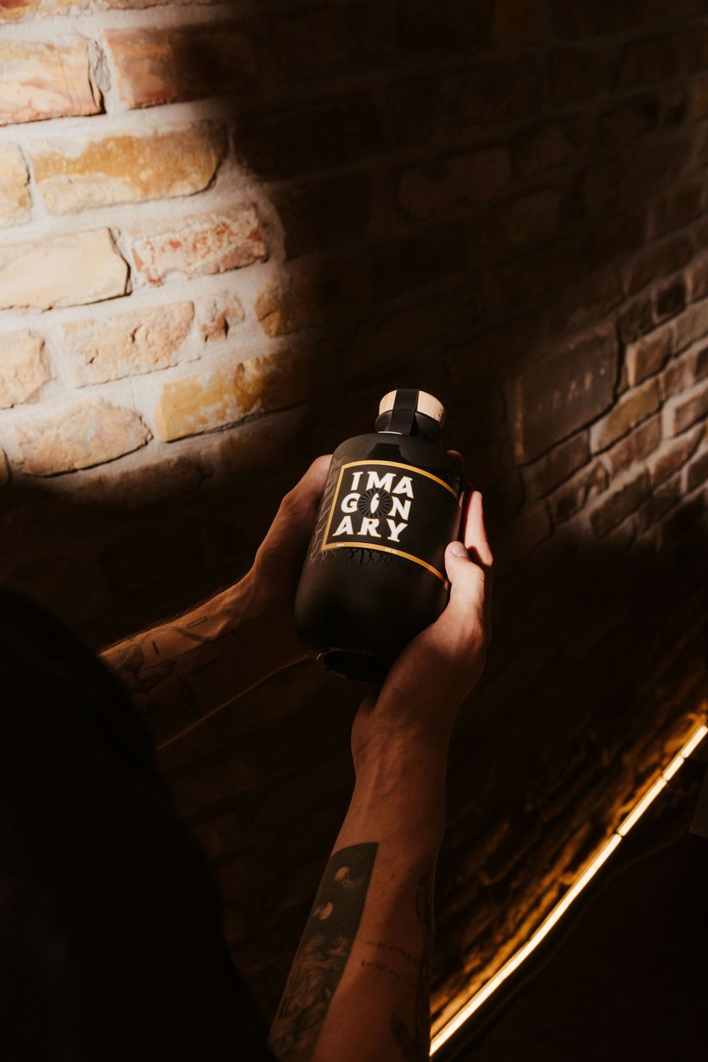 Tattooed hands holding dark bottle labeled 'Imaginary' against brick wall background.