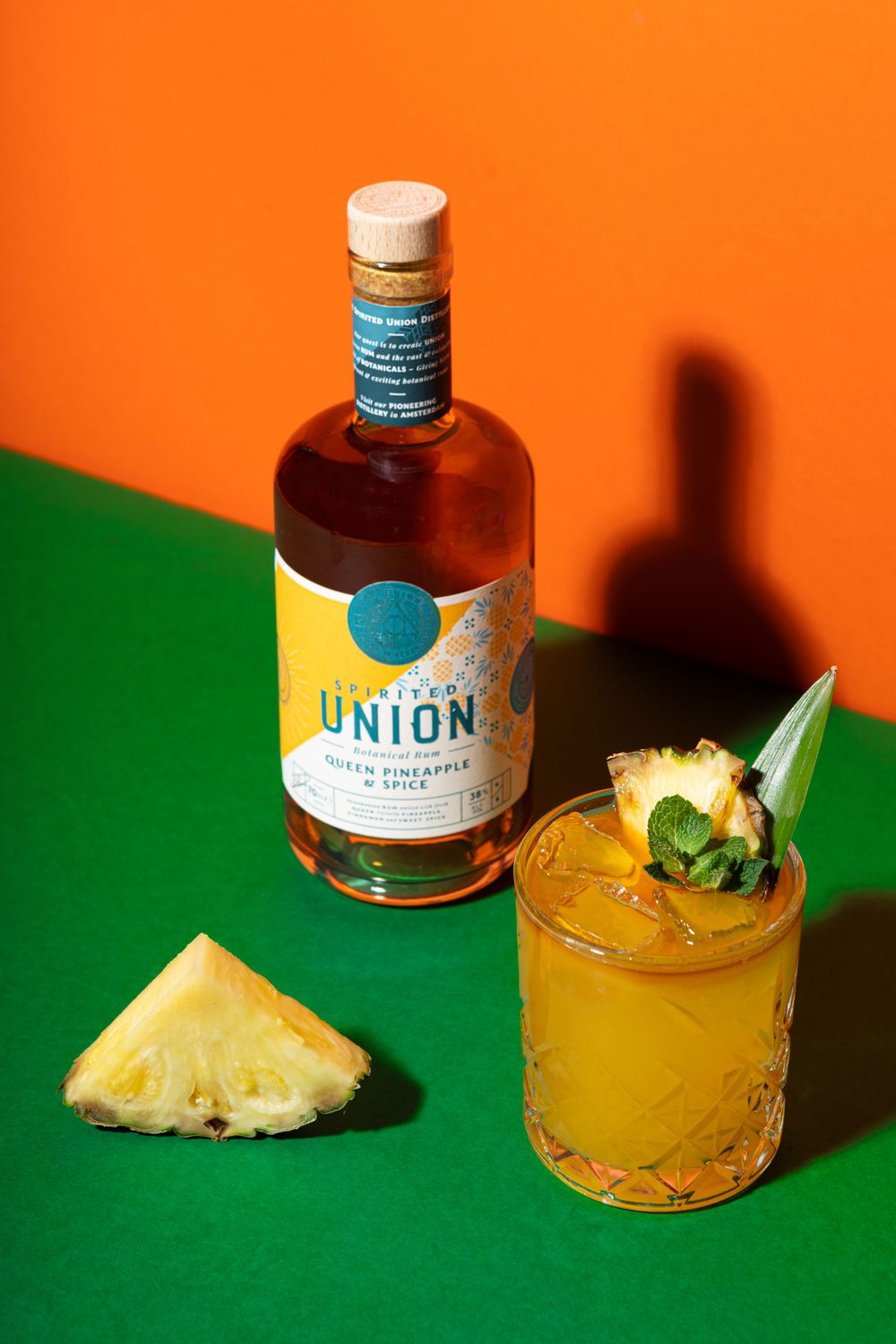 Bottle of Spirited Union liquor with tropical cocktail and pineapple garnish on green background.