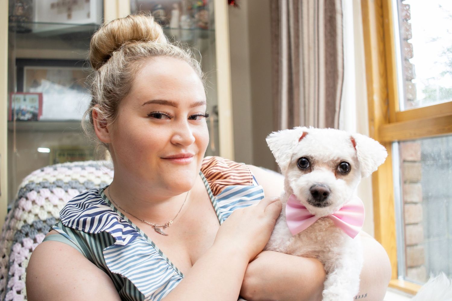 A small white dog wearing a pink bow tie sits next to someone by a sunny window.