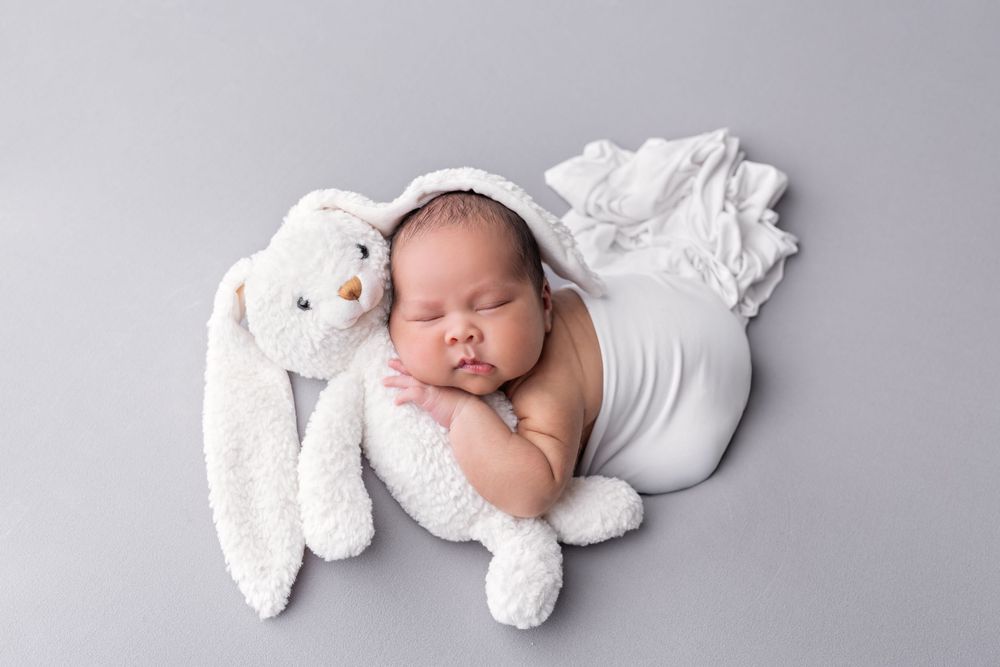Newborn baby sleeping peacefully in white wrap beside a fluffy white bunny stuffed animal on gray background.