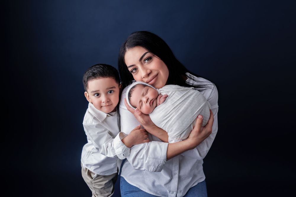 A mother smiles warmly holding a newborn and embracing her young son against a dark blue studio background.