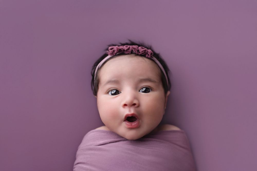 Newborn baby girl wrapped in purple swaddle with floral headband, making a surprised expression on purple background.