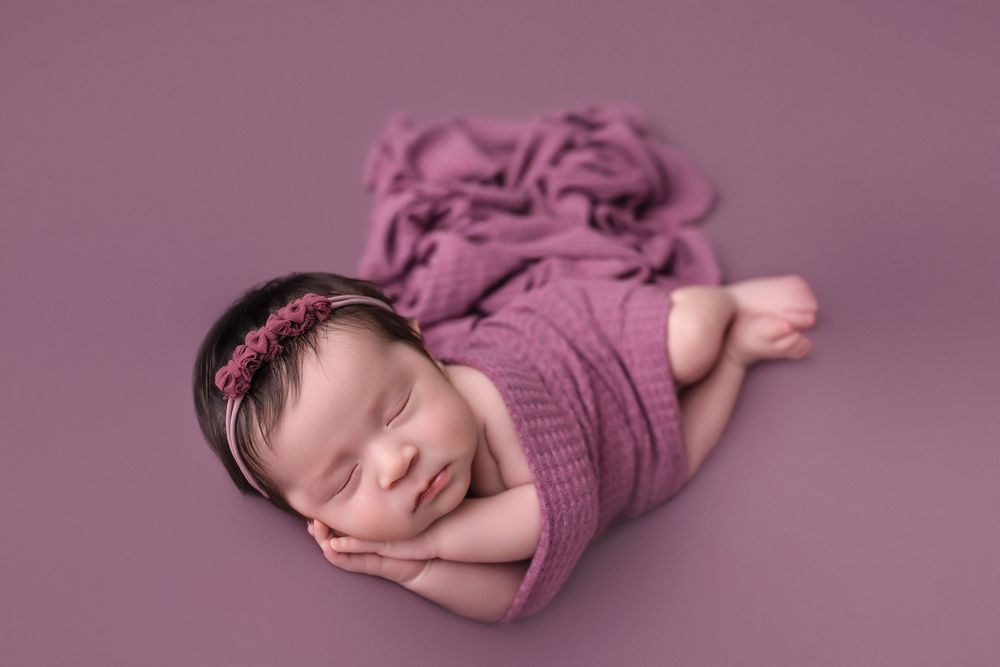 Sleeping newborn baby girl wrapped in a pink mauve knit swaddle with a floral headband on a matching background.