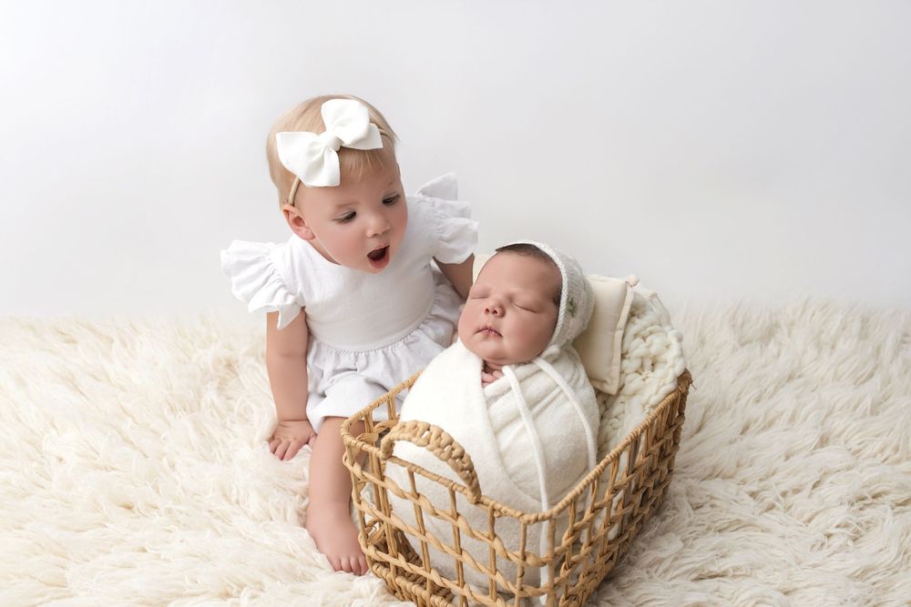 Two babies in white outfits sit together on a fluffy rug, one in a wicker basket wearing a bonnet.
