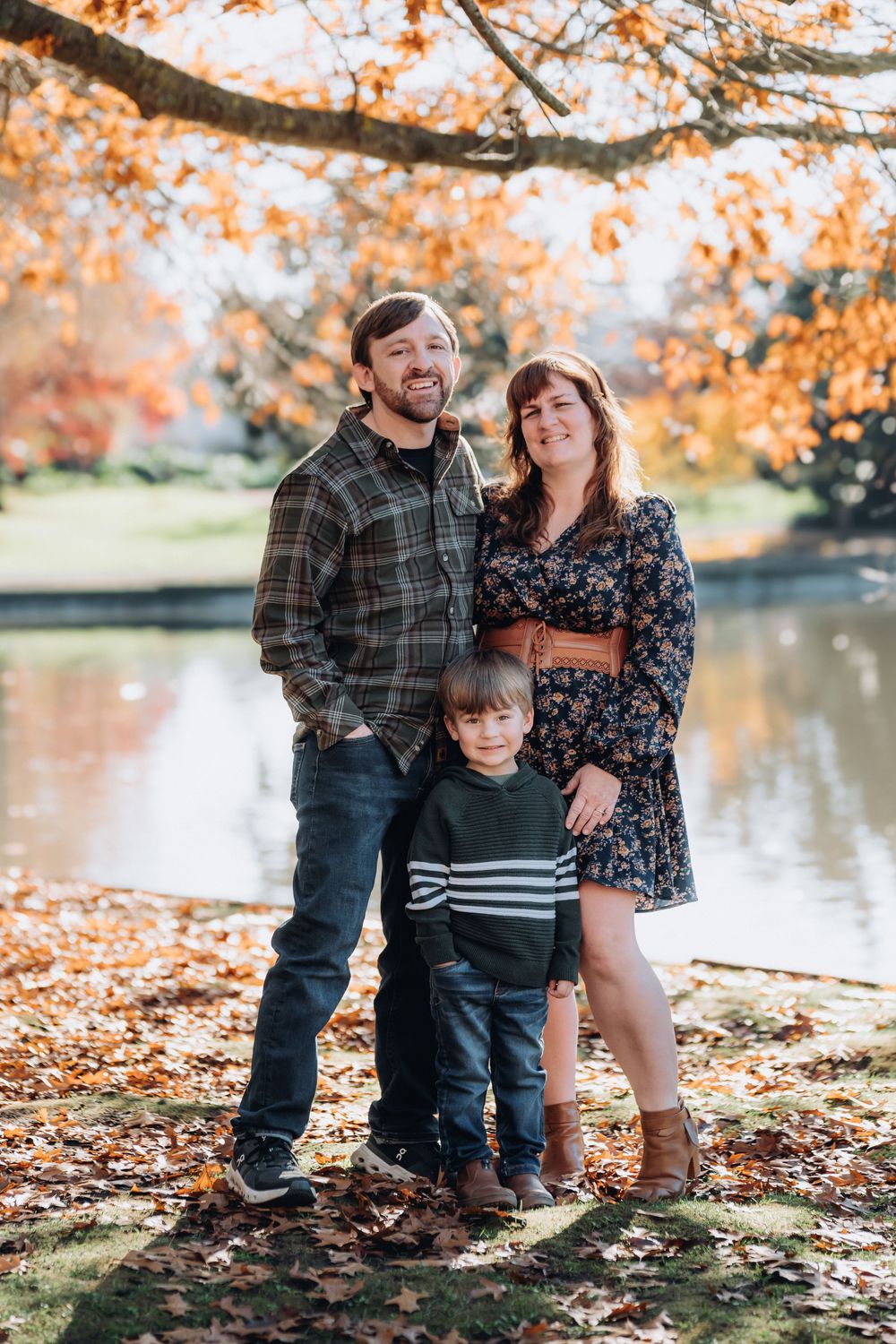 Family of three posing by a pond surrounded by vibrant autumn foliage.