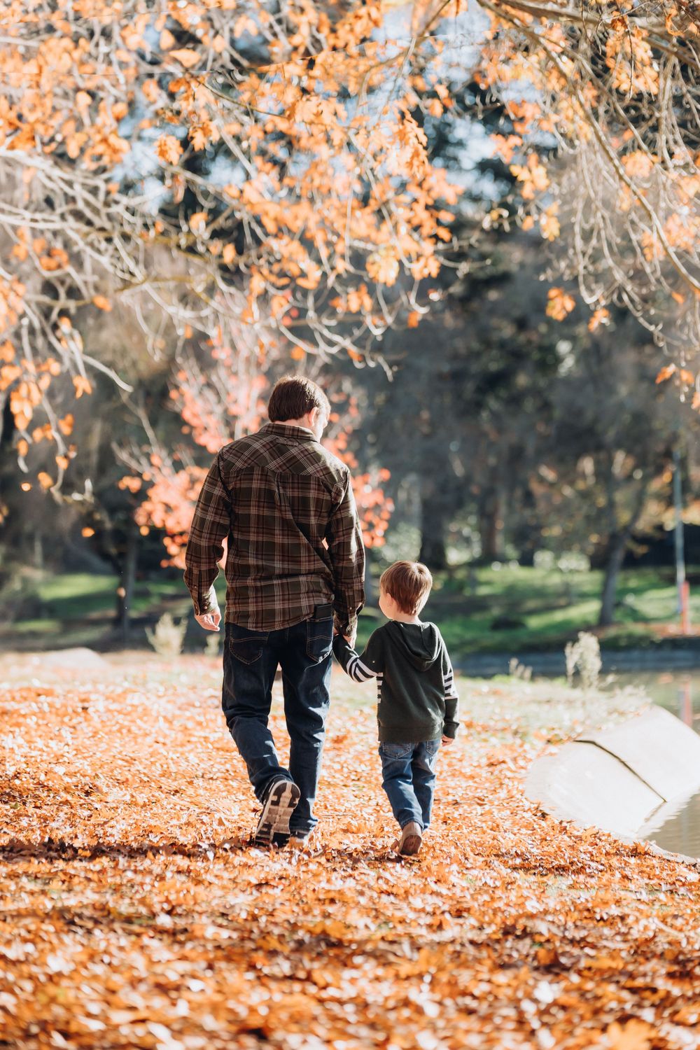 Father and son walking hand in hand through a park covered in fall leaves.